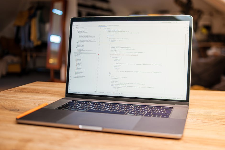 The image shows a slim, modern laptop with a large screen displaying lines of code or a text editor, positioned on a smooth wooden table. The keyboard appears backlit and is black with white character